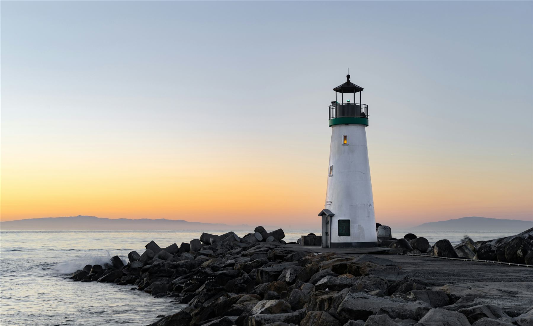 A white lighthouse on a rocky breakwater at sunset, waves breaking against the rocks.